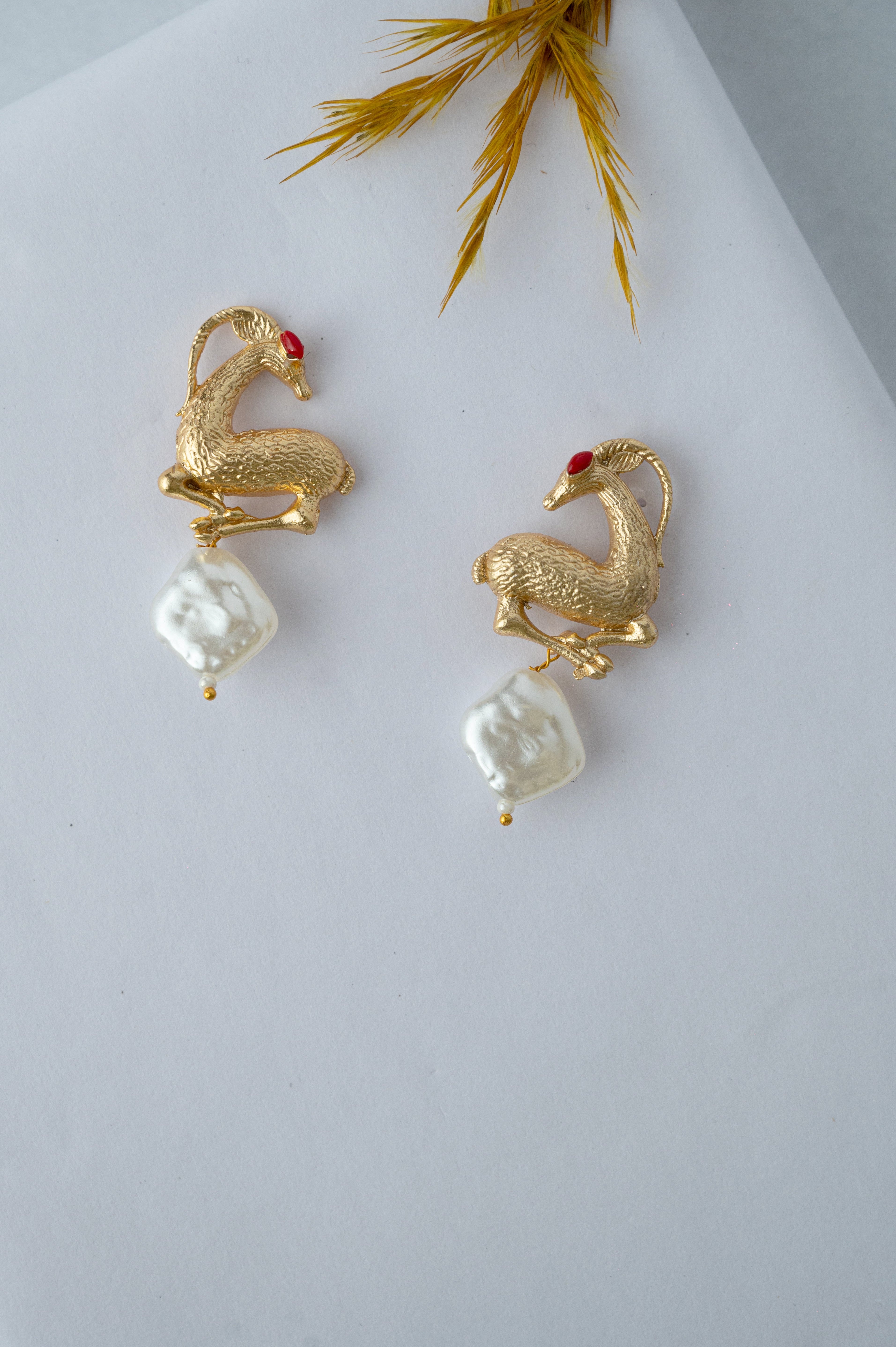 Pair of gold and pearl earrings on a light gray background with dried grass at the bottom.
